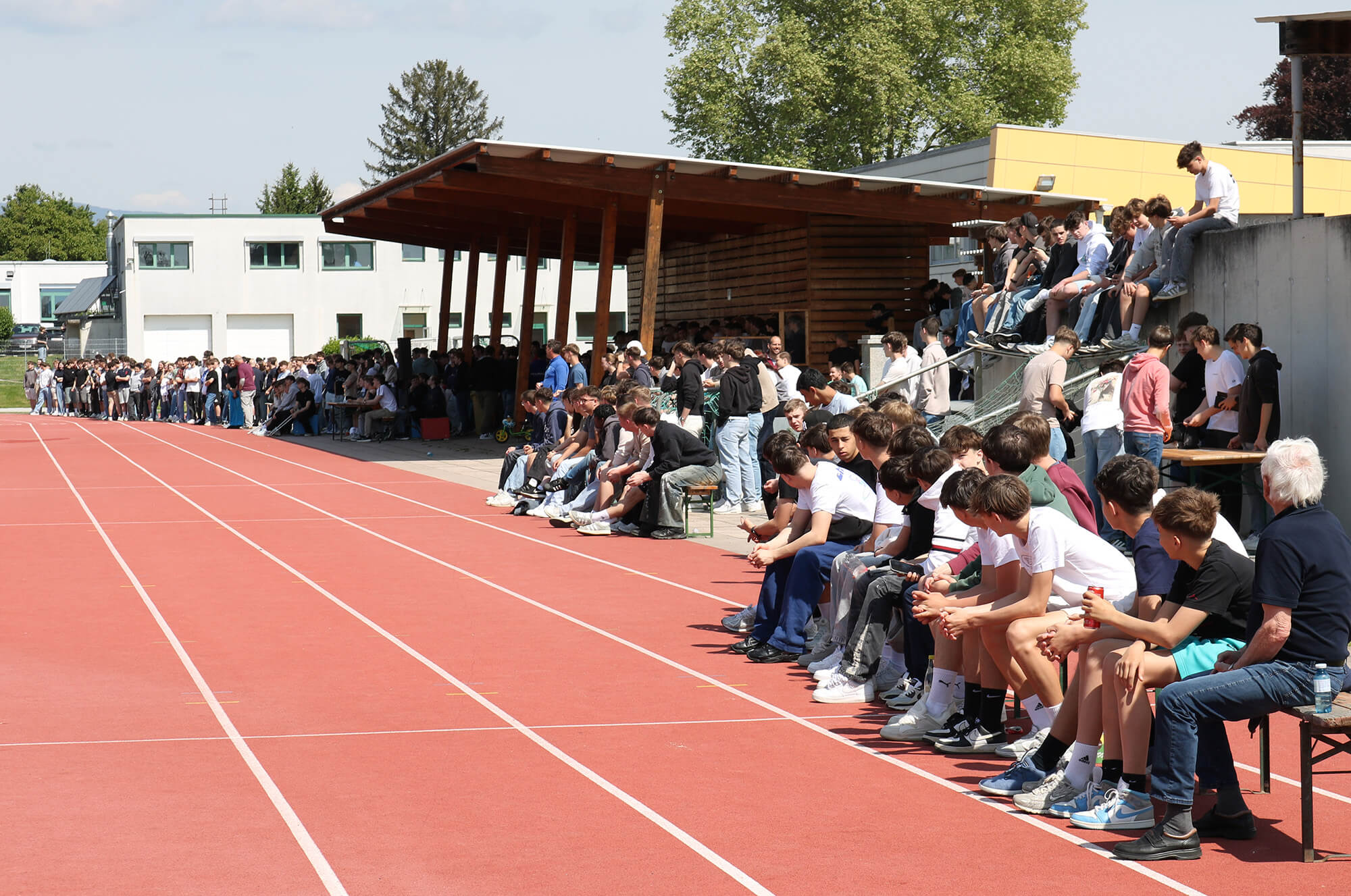 Gruppe von Menschen sitzt und steht auf Tribüne neben einer Leichtathletikbahn bei sonnigem Wetter, Zuschauer im Freien.