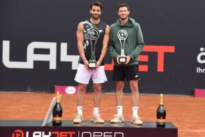 Zwei Tennisspieler mit Trophäen auf dem Siegerpodest der Layjet Open, feiernd auf rotem Sandplatz.