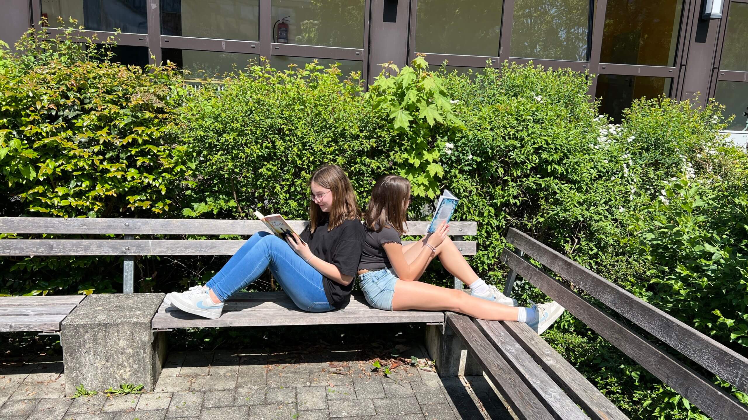 Zwei Mädchen genießen die Sonne auf einer Parkbank und lesen draußen Bücher im Sommer.