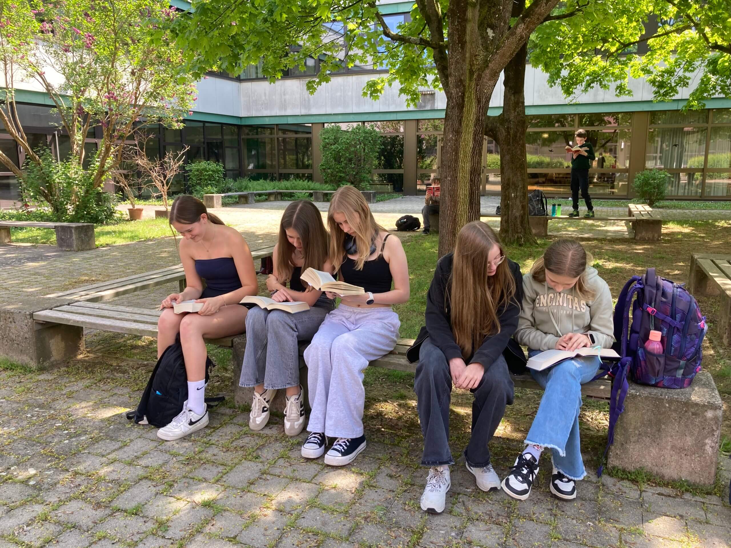 Schülergruppe liest im sonnigen Schulgarten, vertieft in Bücher, umgeben von grüner Natur und Rucksäcken.