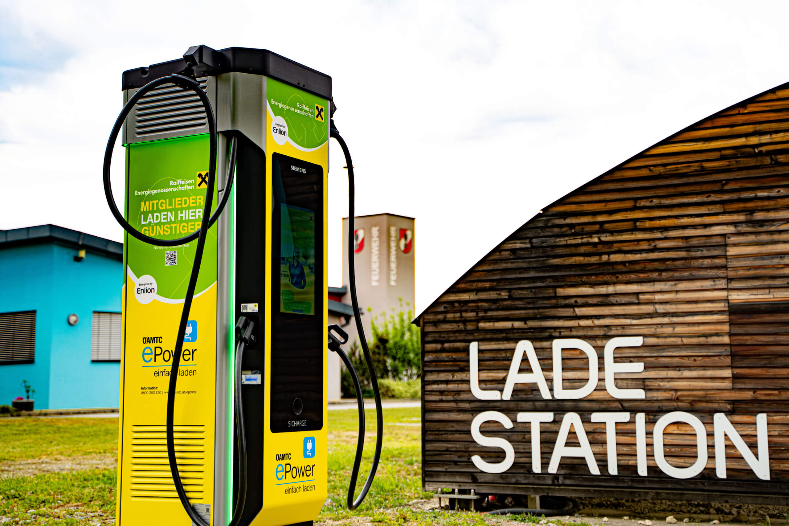 E-Ladestation im Freien mit grüner Beschilderung und Holzgebäude im Hintergrund, ideale Aufladung für Mitglieder.