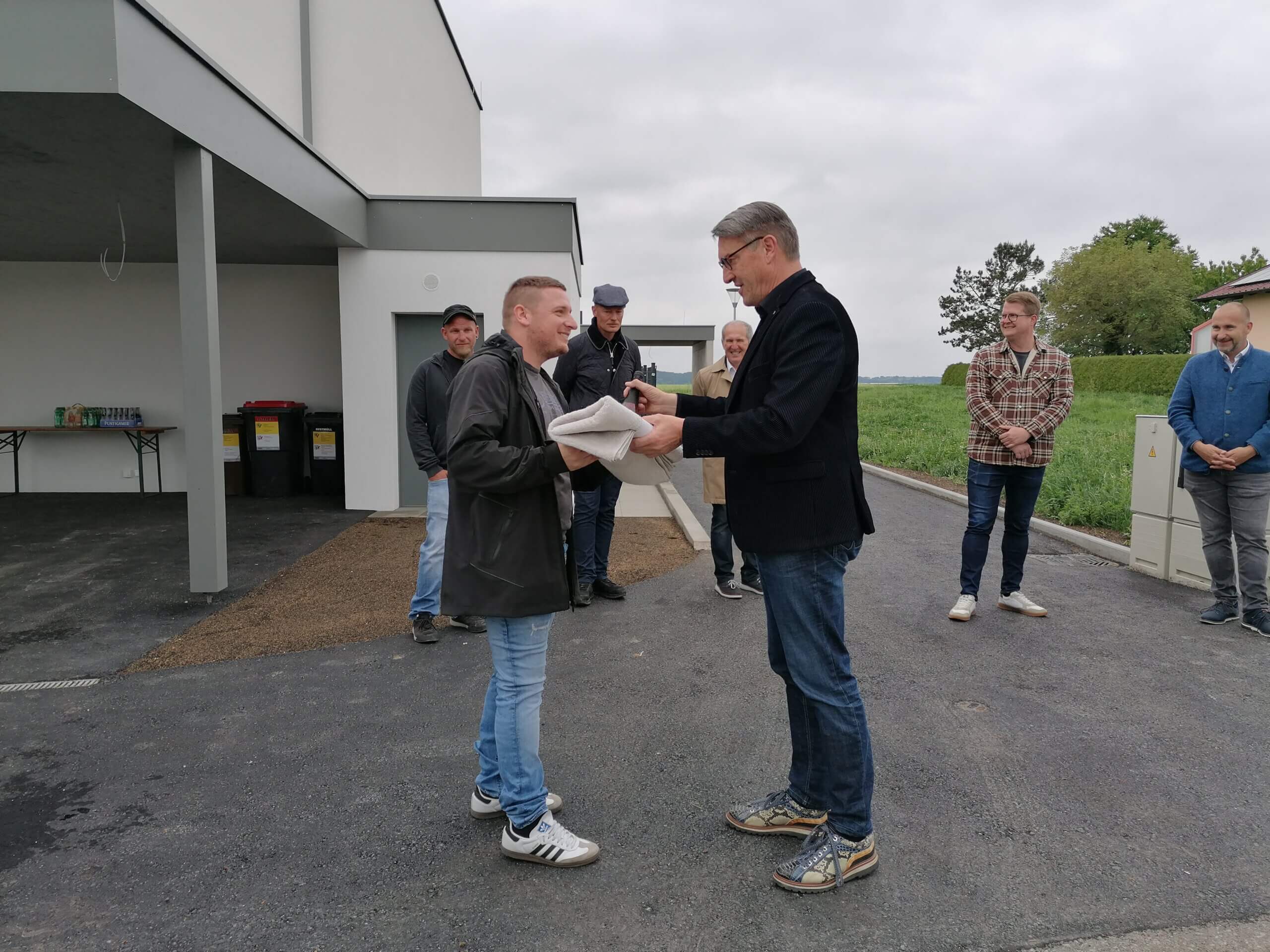 Person erhält feierlich ein Geschenk im Freien, umgeben von einer Gruppe gut gelaunter Menschen vor einem Gebäude.