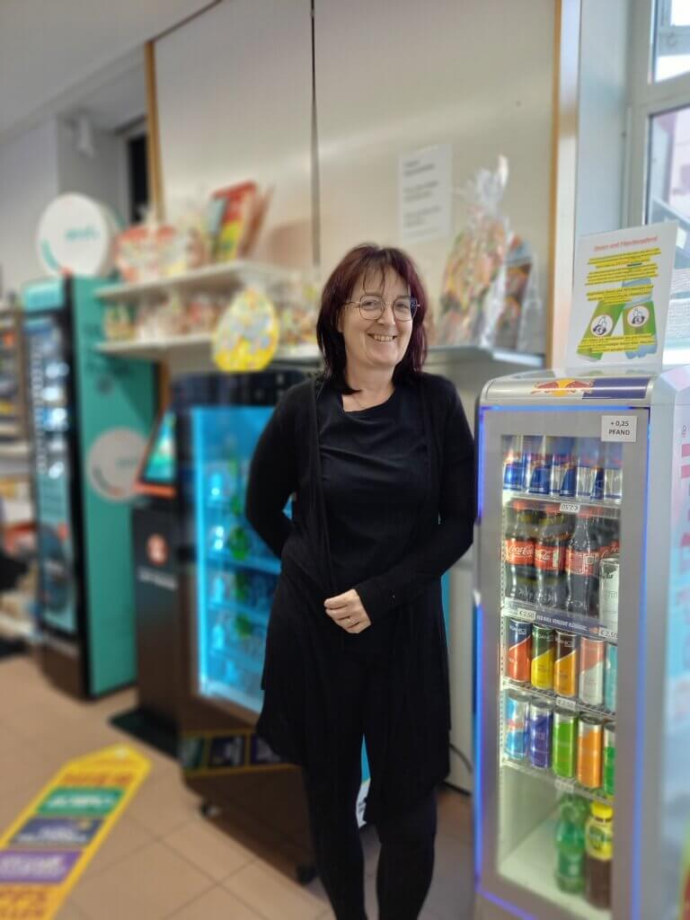 Trafik Wuggenig_Romana Brandstätter2©Nicole Matsch Frau im Geschäft vor Kühlschrank mit Getränken, Snacks im Hintergrund, lächelnd.