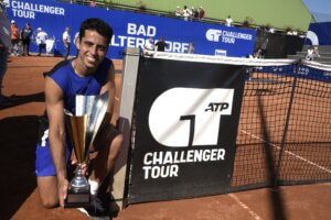 Tennisspieler mit Pokal beim ATP Challenger Tour in Bad Waltersdorf, lächelnd neben Tennisnetz auf rotem Sandplatz.
