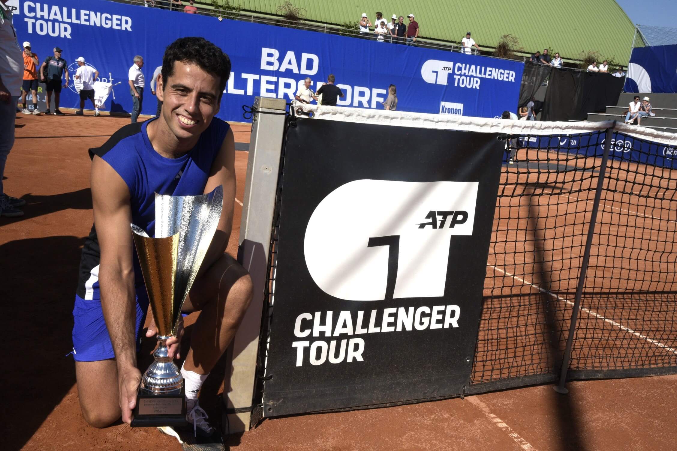Tennisspieler mit Pokal beim ATP Challenger Tour in Bad Waltersdorf, lächelnd neben Tennisnetz auf rotem Sandplatz.
