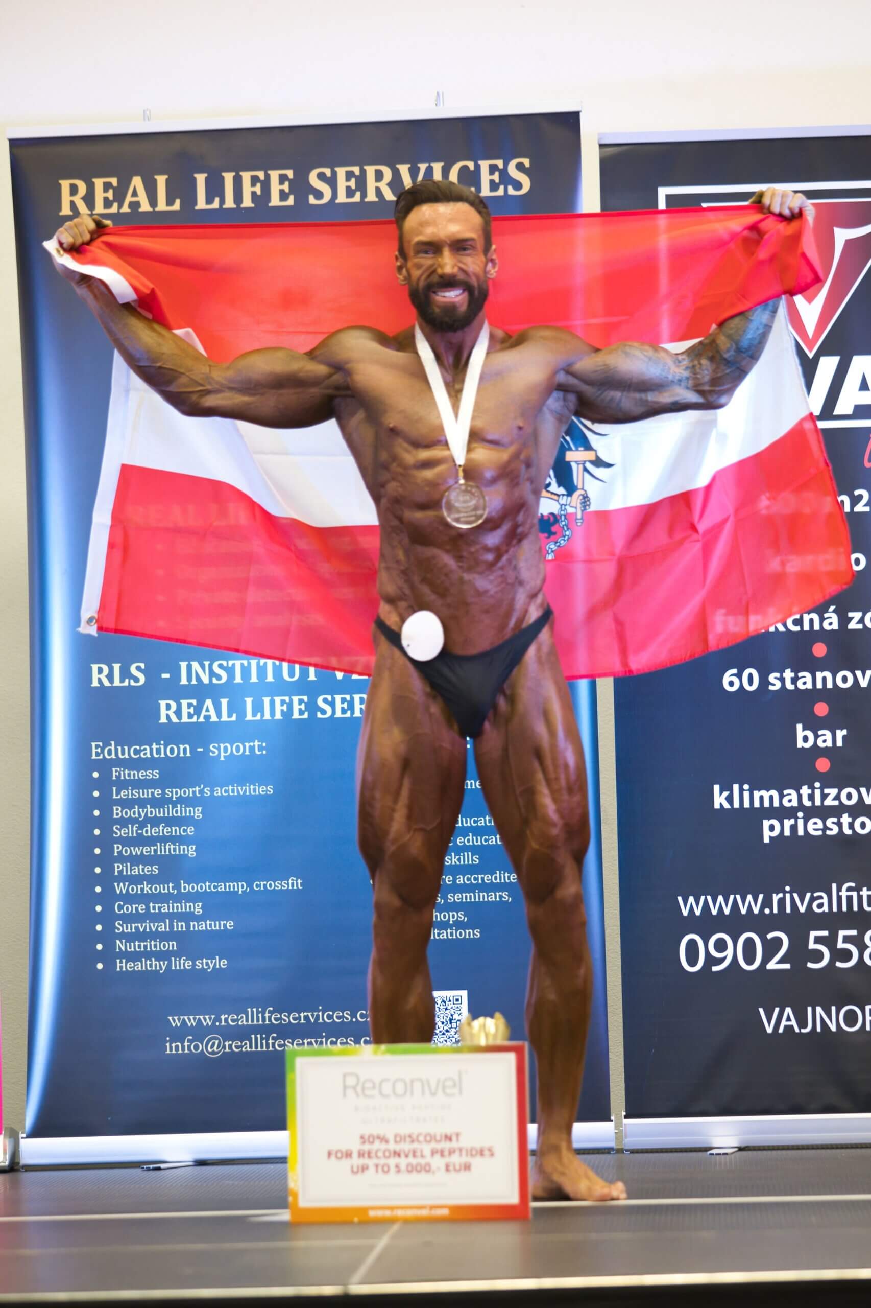 Bodybuilder präsentiert Flagge bei Fitness-Wettbewerb, mit Medaille und Werbeangebot im Hintergrund.