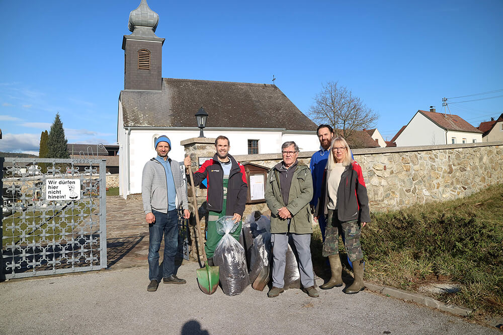 Gemeinschaftsarbeiten vor Dorfkirche: Fünf Personen mit Schaufeln und Gartensäcken bei sonnigem Wetter.