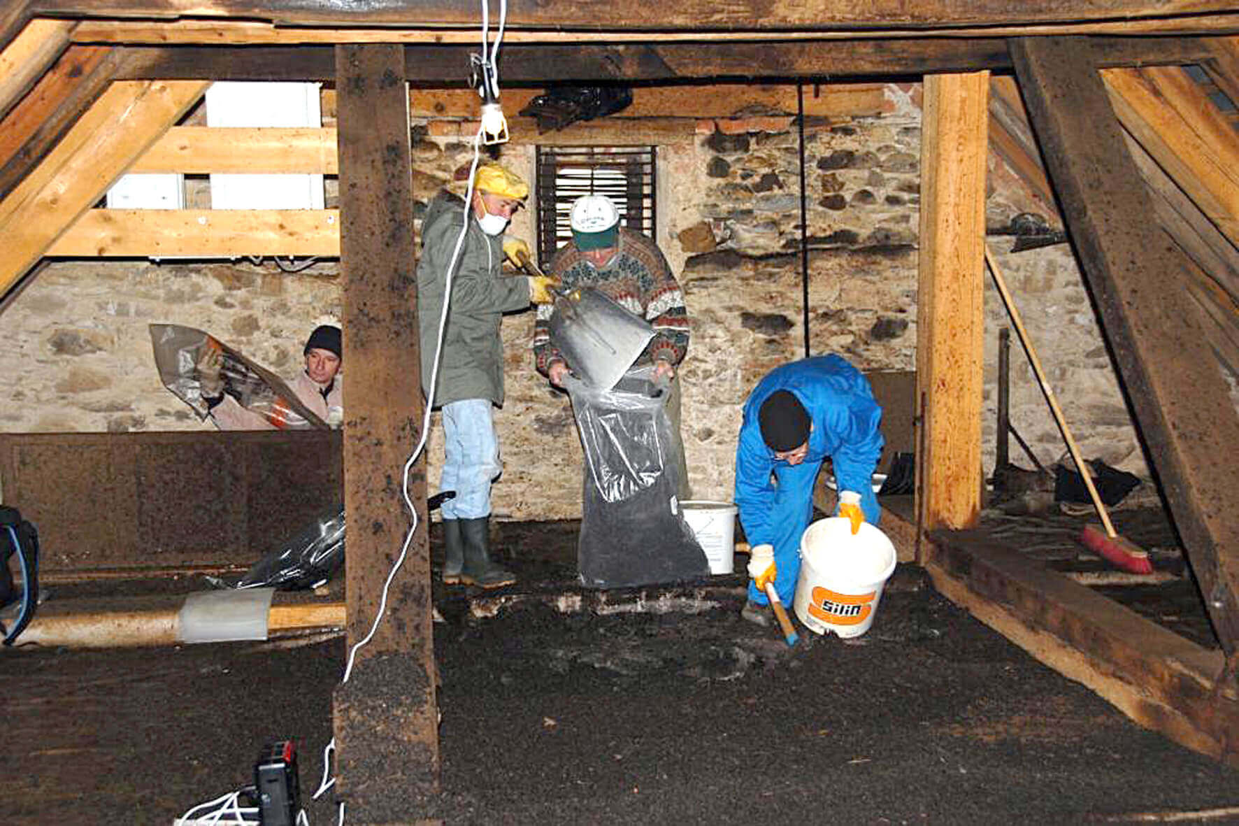 Arbeiter in einem Dachboden führen Sanierungsarbeiten durch, trennen und verpacken altes Material sicher in Säcke.