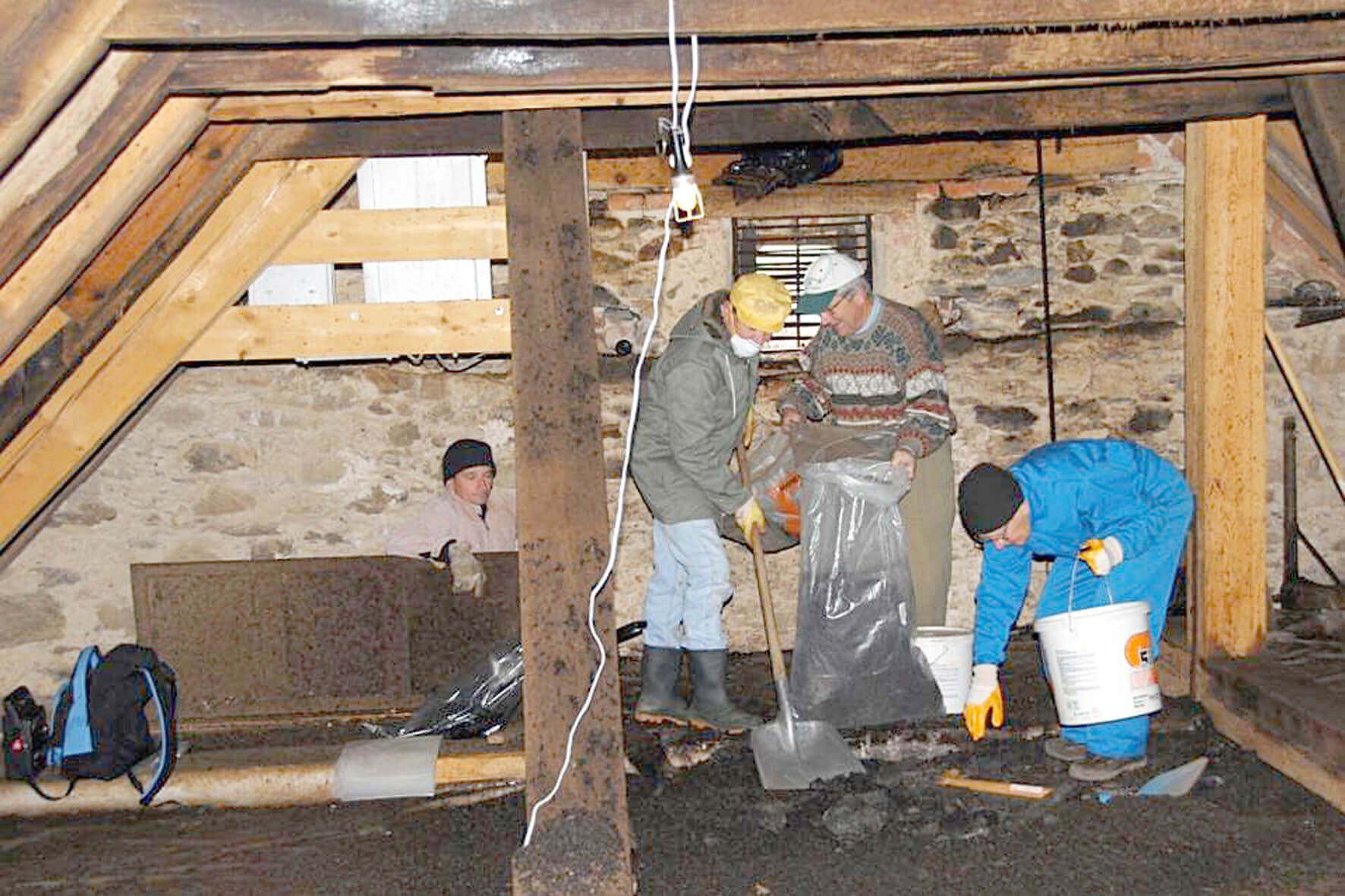 Menschen reinigen Dachboden mit Schaufel und Eimern, tragen Schutzkleidung in rustikalem Gebäude. Renovierung im Gange.