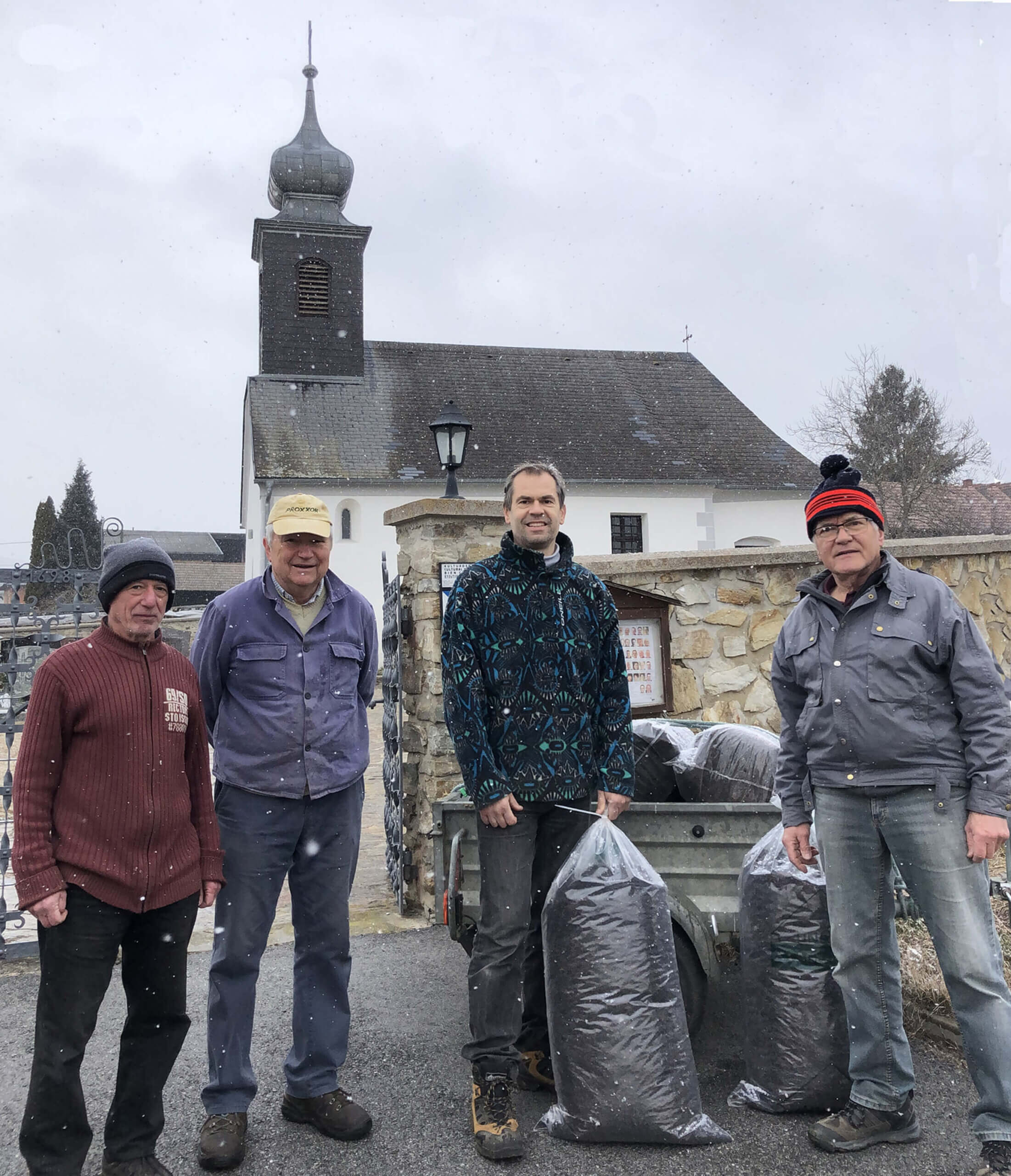 Vier Männer stehen vor einer Kirche mit Säcken voller Erde im Schnee. Gebäudekirchenglocke im Hintergrund.