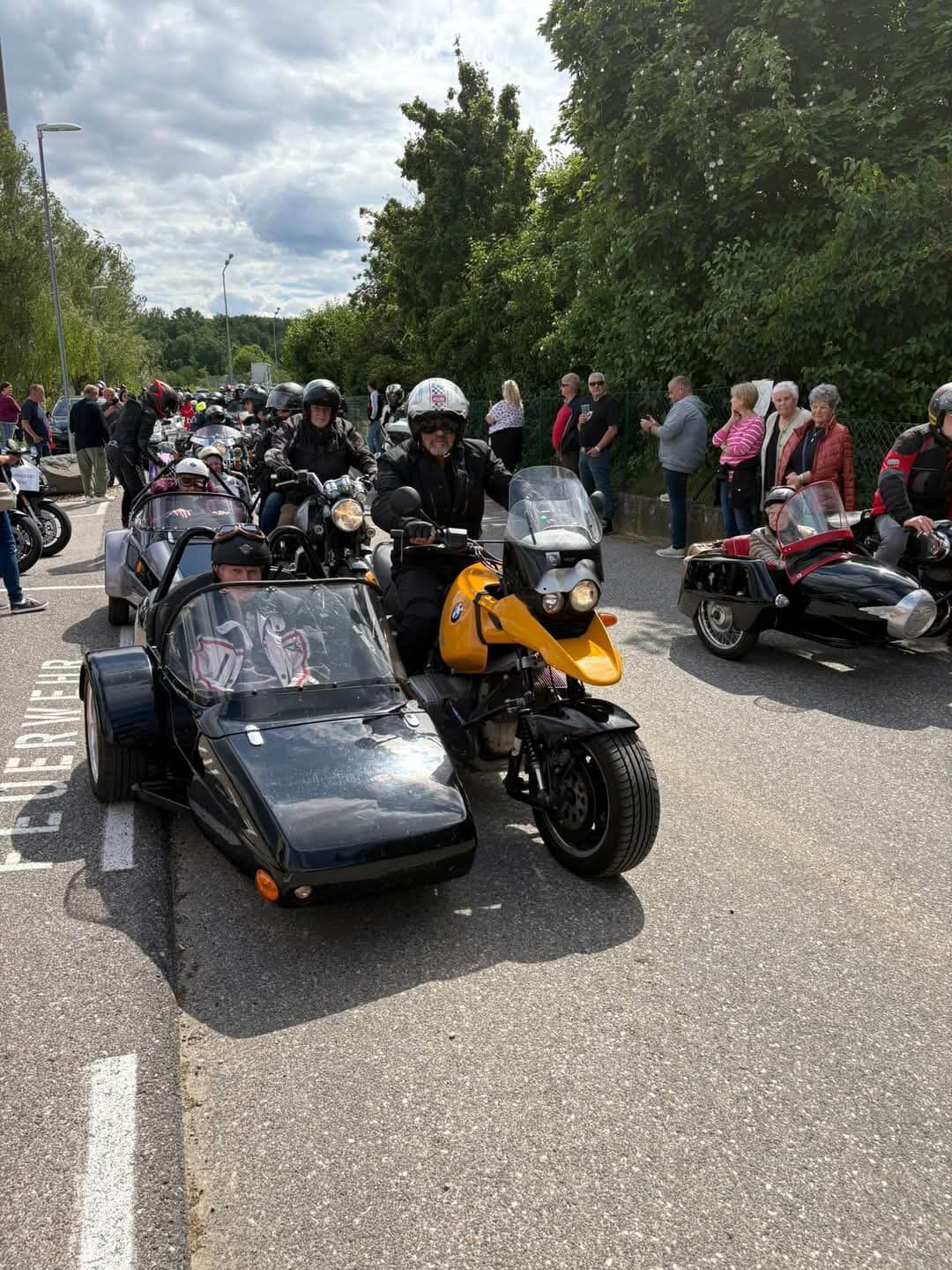 Motorradtreffen mit Beiwagen auf Straße, umgeben von Zuschauern und grüner Landschaft unter bewölktem Himmel.