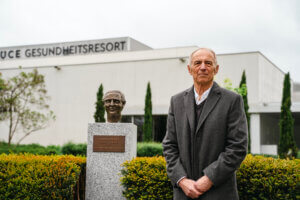 Älterer Mann neben Büste und Schild vor dem Ruce Gesundheitsresort, umgeben von Pflanzen und moderner Architektur.