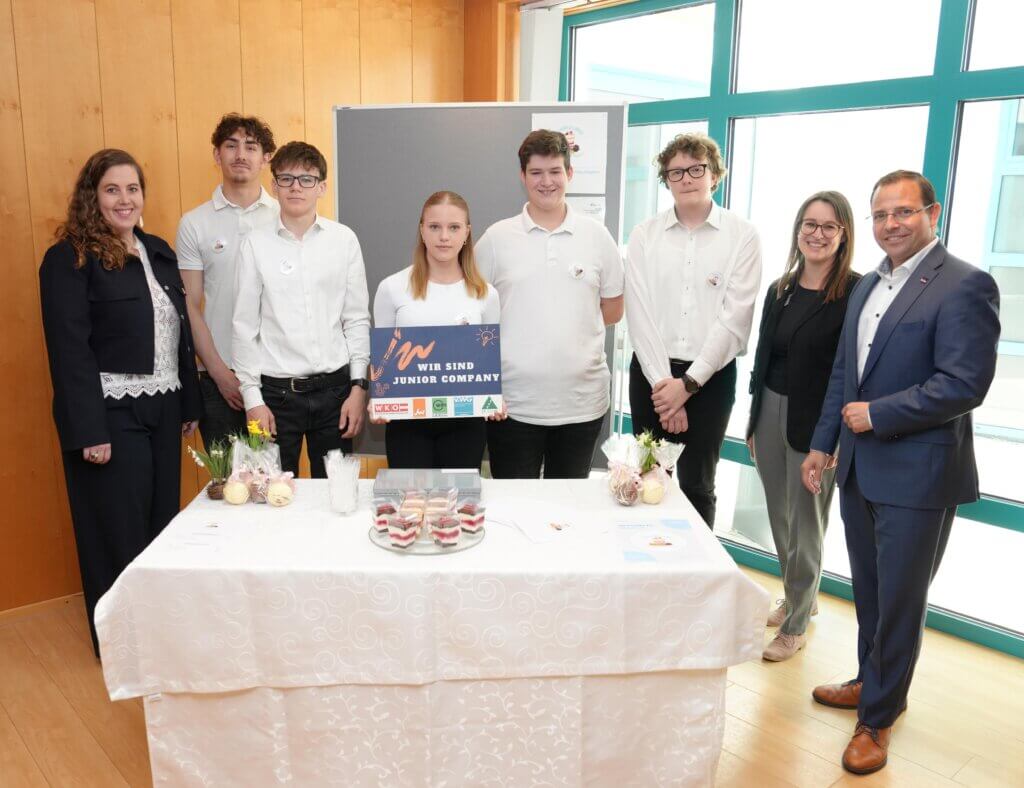 Gruppe von Schülern präsentiert ihre Junior Company auf einer Messe mit Unterstützung von Lehrkräften.