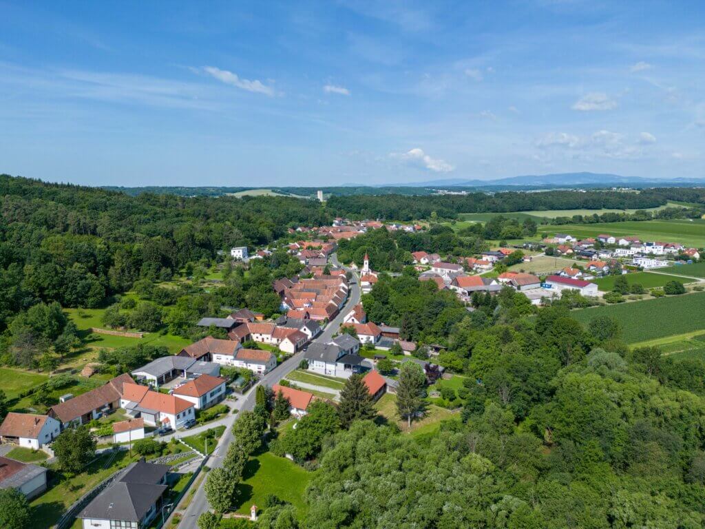 Luftaufnahme eines idyllischen Dorfes mit Kirche, umgeben von Wäldern und Feldern unter blauem Himmel.