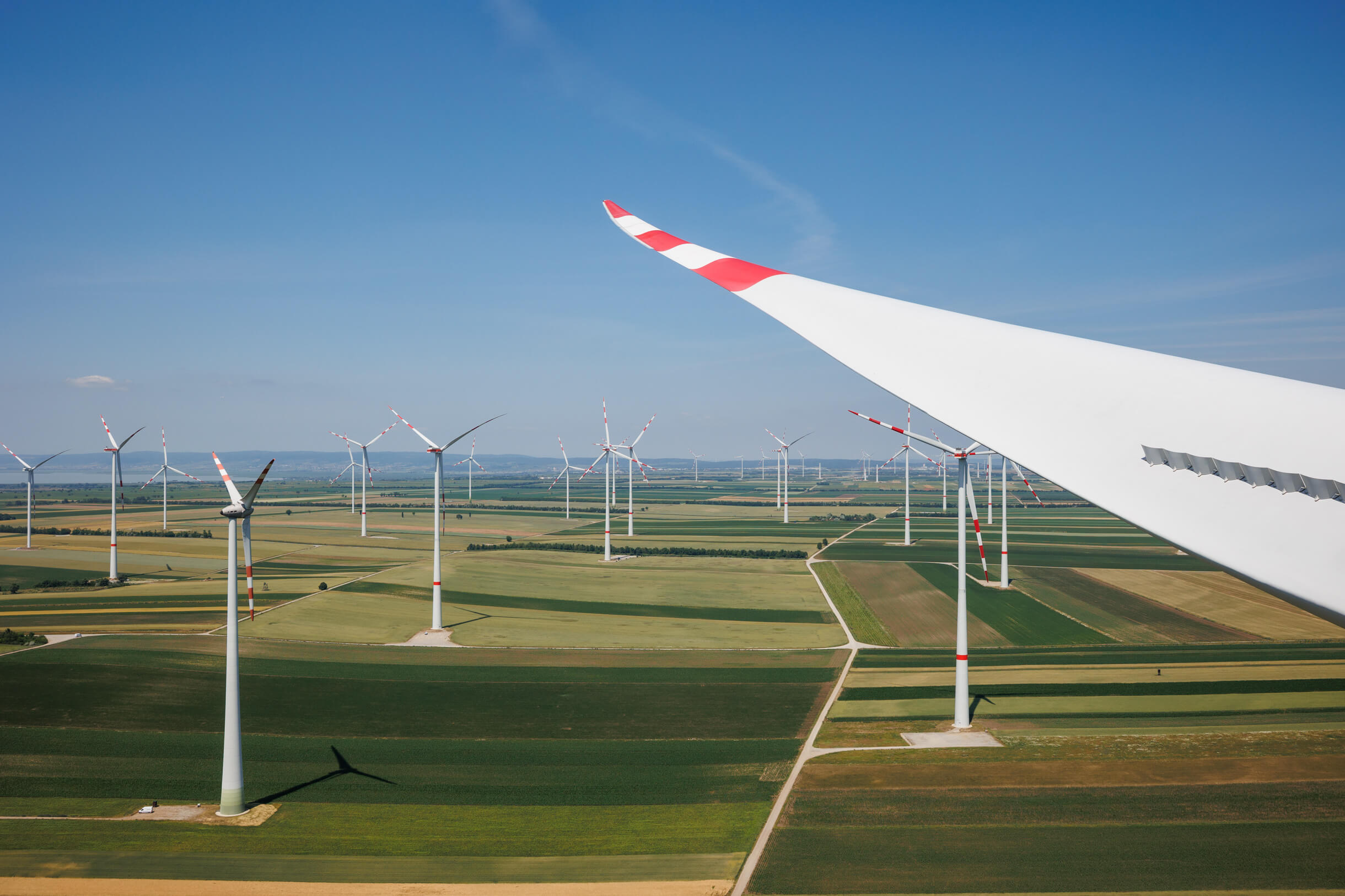 Windkraftanlage in Landschaft, großes Rotorblatt im Vordergrund, mehrere Turbinen im Hintergrund unter blauem Himmel.