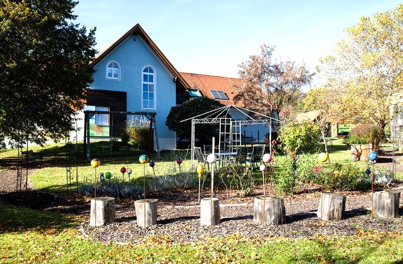 Ruhiger Garten mit dekorativen Kugeln, Pavillon und blauem Haus im Hintergrund an einem sonnigen Herbsttag.