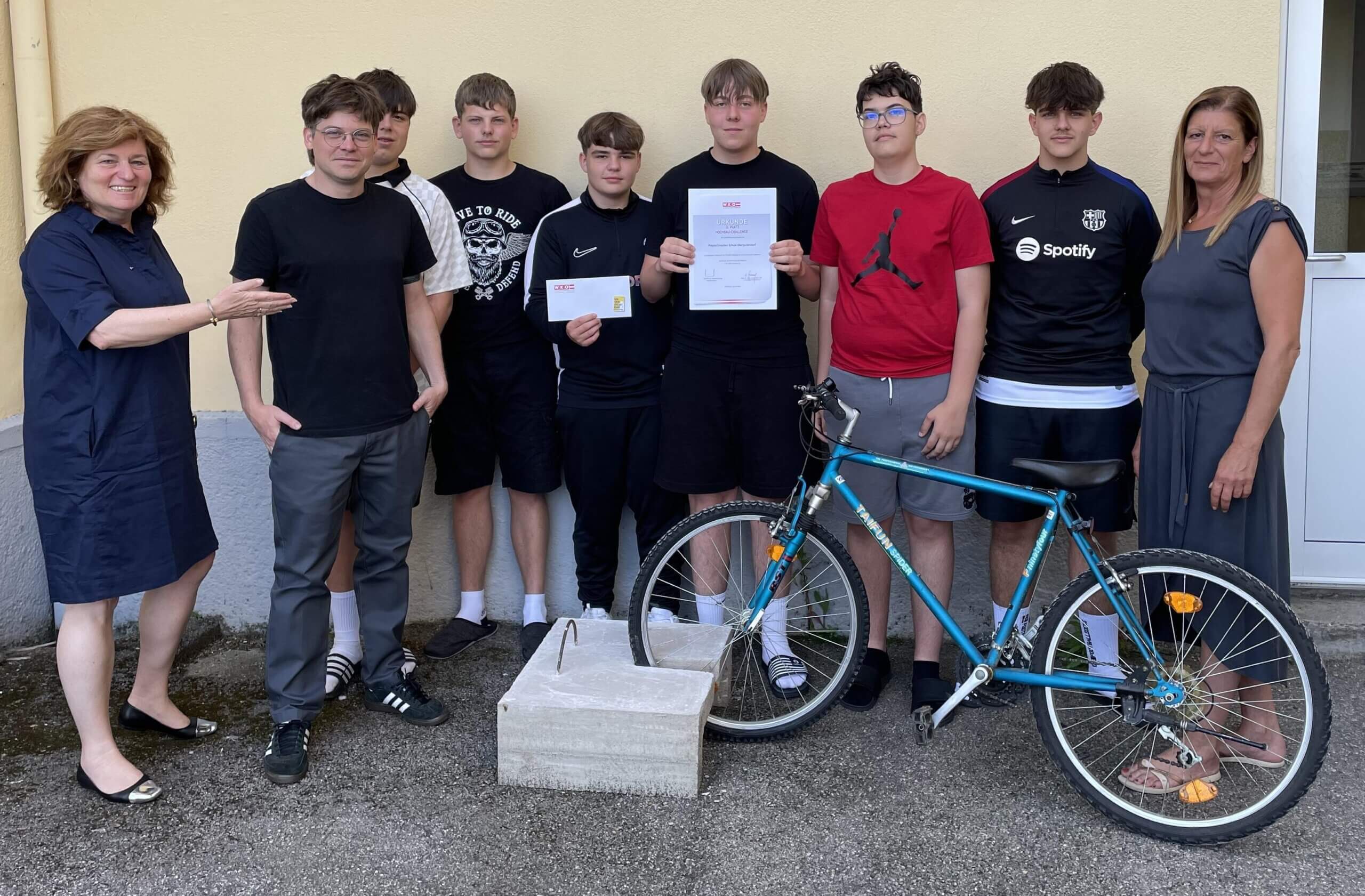 Gruppe junger Menschen mit Zertifikaten und Fahrrad bei Preisverleihung vor gelber Wand.