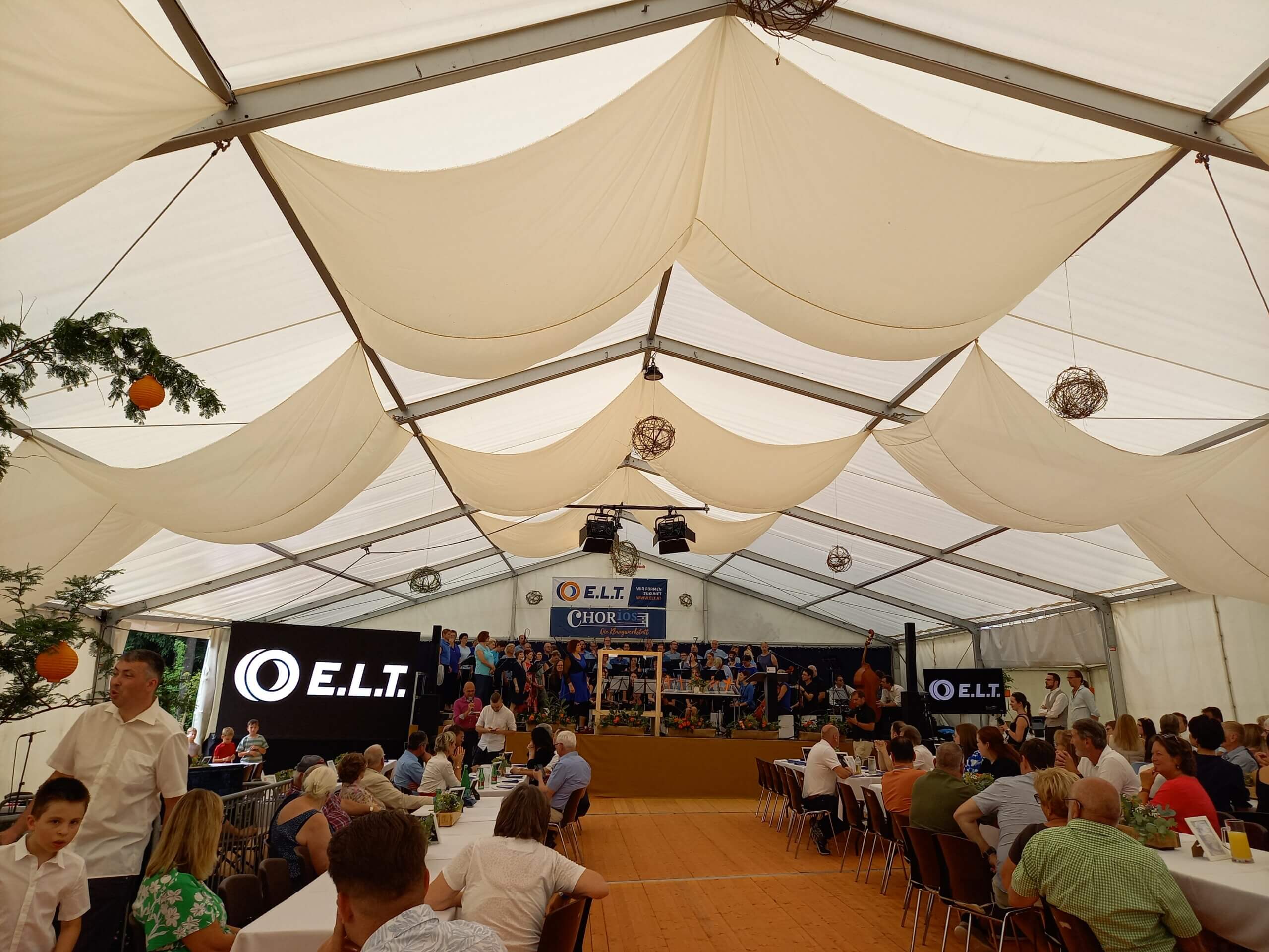 Festzelt mit Chorauftritt auf der Bühne, Publikum an Tischen, Dekoration und E.L.T.-Logo auf Bildschirmen.