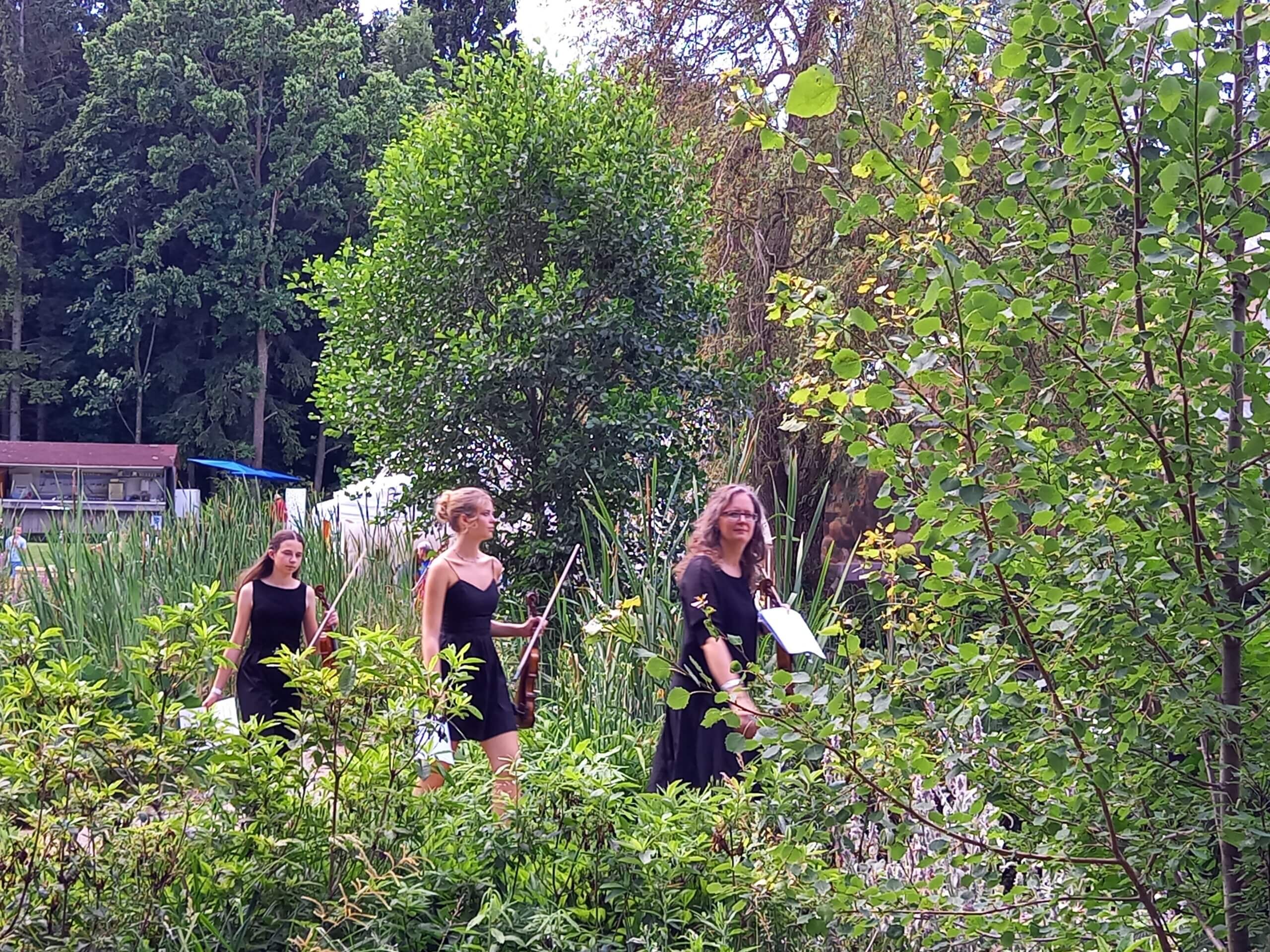 Frauen in schwarzen Kleidern mit Violinen spazieren durch einen grünen Garten mit Bäumen und Büschen.