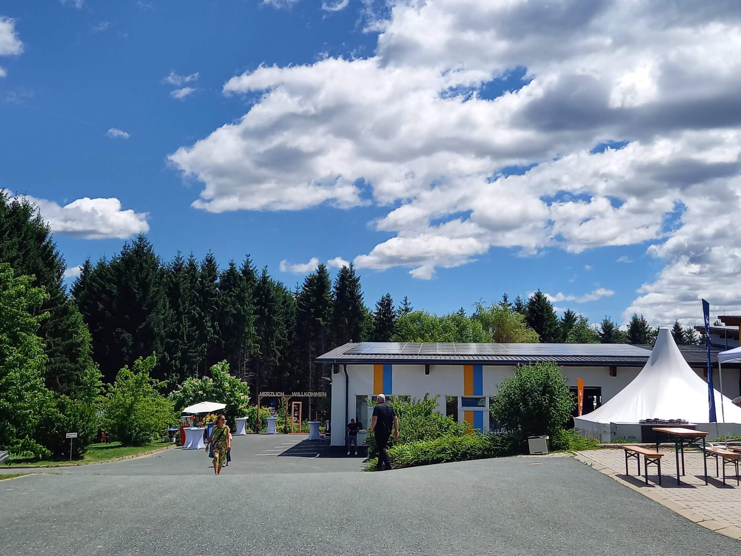 Gebäude mit Solardach, Menschen beim Sommerfest, Zelte und Bäume unter blauem Himmel mit Wolken.