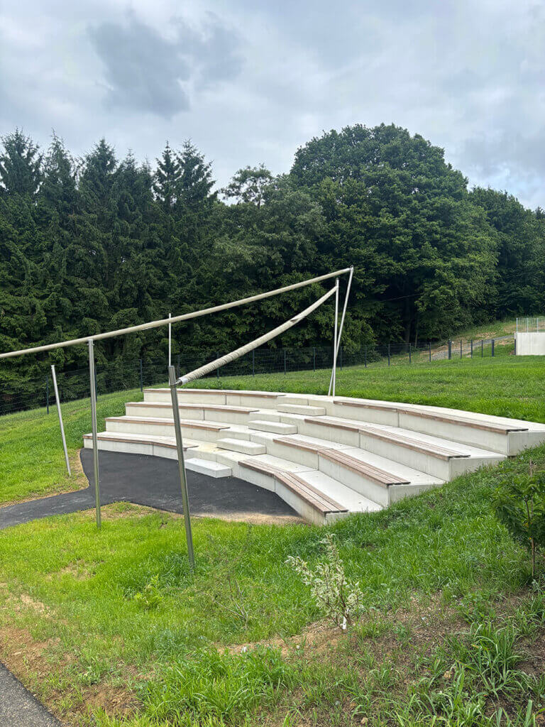4©Chiara Pieler Moderne Betontribüne im Freien, umgeben von grüner Natur und Bäumen an einem bewölkten Tag.
