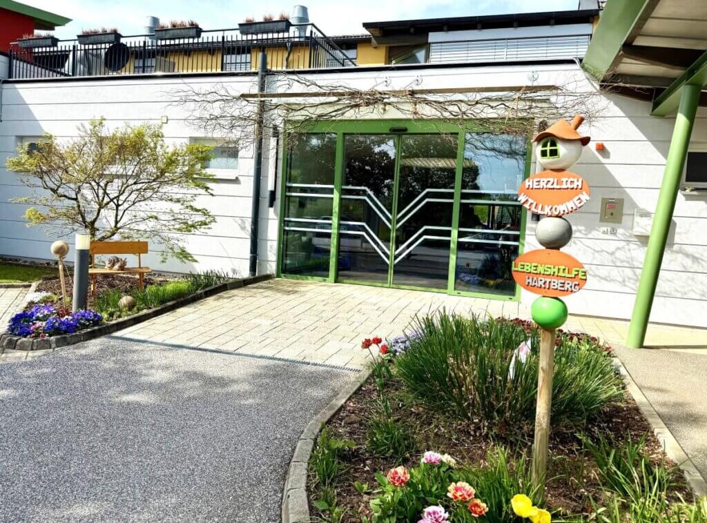 Eingang Lebenshilfe Hartberg, Österreich: Begrüßungsschild mit Blumenbeet und moderner Glasfassade im Sonnenschein.