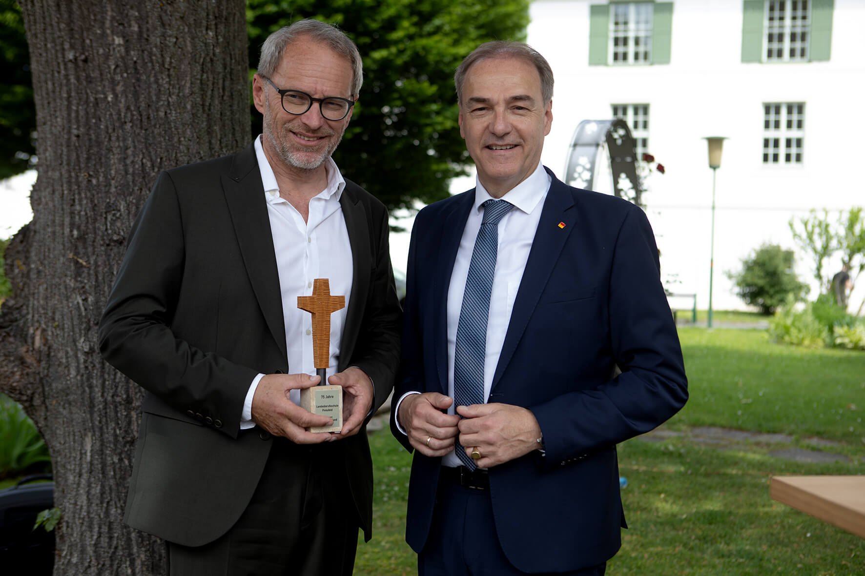 Zwei Männer in Anzügen, einer hält eine Auszeichnung in Form eines Kreuzes, posieren im Freien vor einem Gebäude.