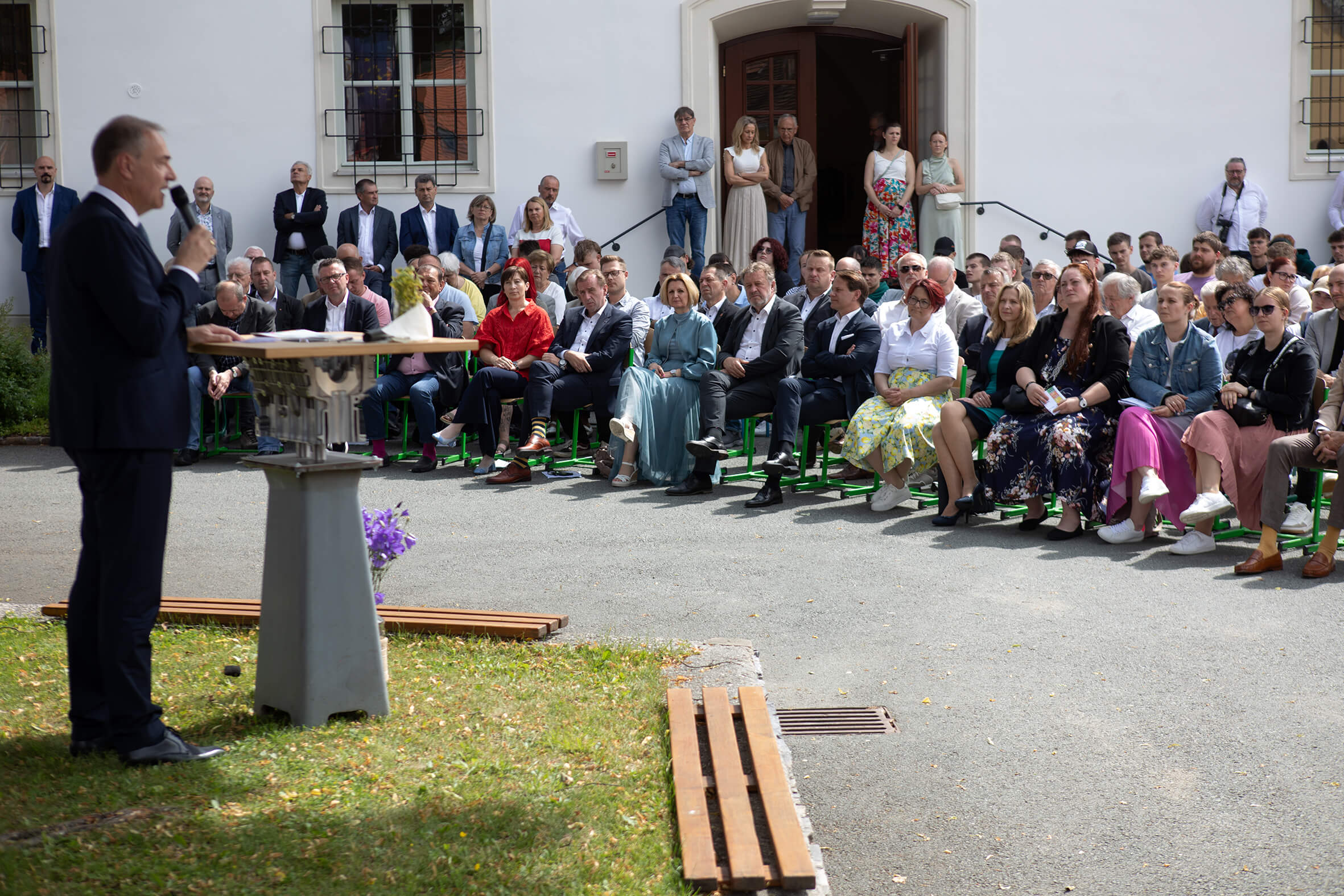 Person hält Rede vor einer sitzenden Gruppe bei einer Outdoor-Veranstaltung. Publikum hört aufmerksam zu.