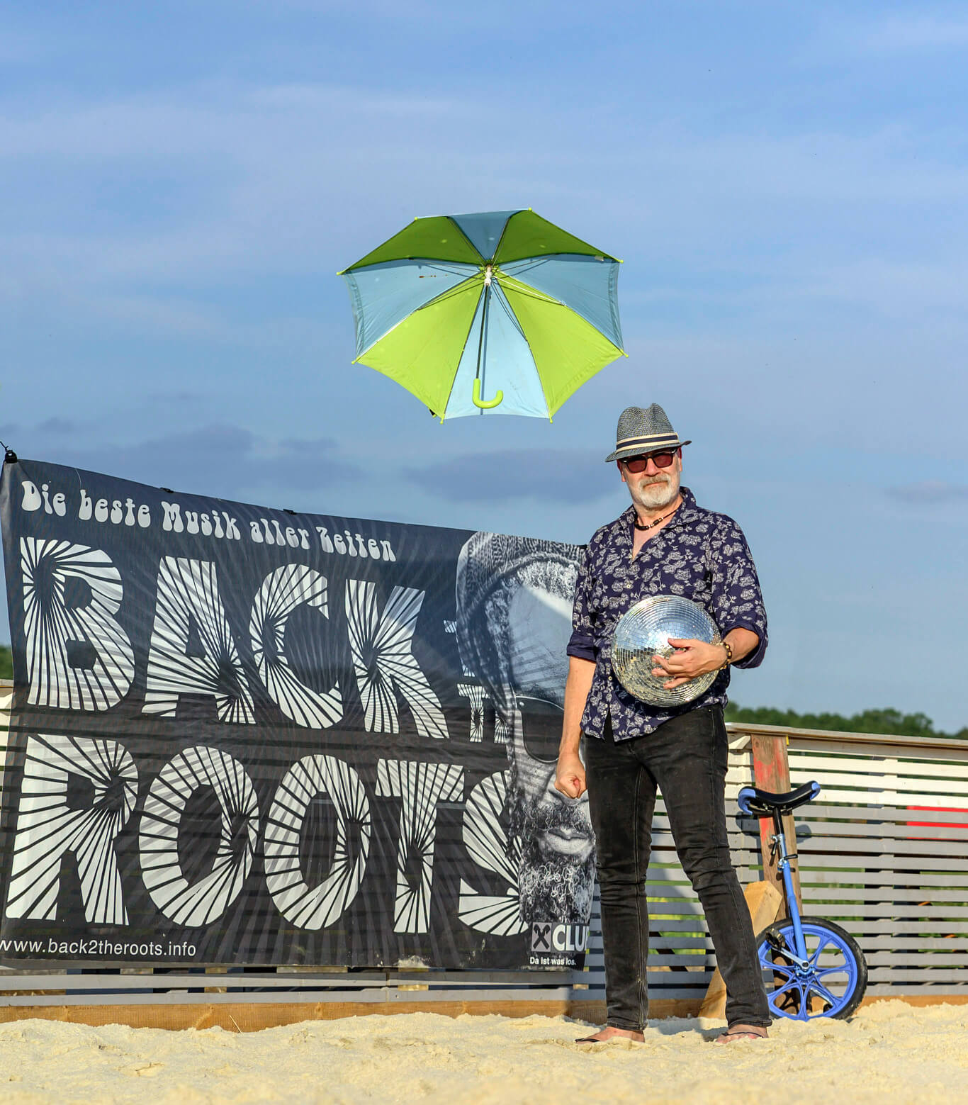 Mann steht vor Back to the Roots-Banner, hält Discokugel, im Hintergrund schwebender Regenschirm und Einrad.