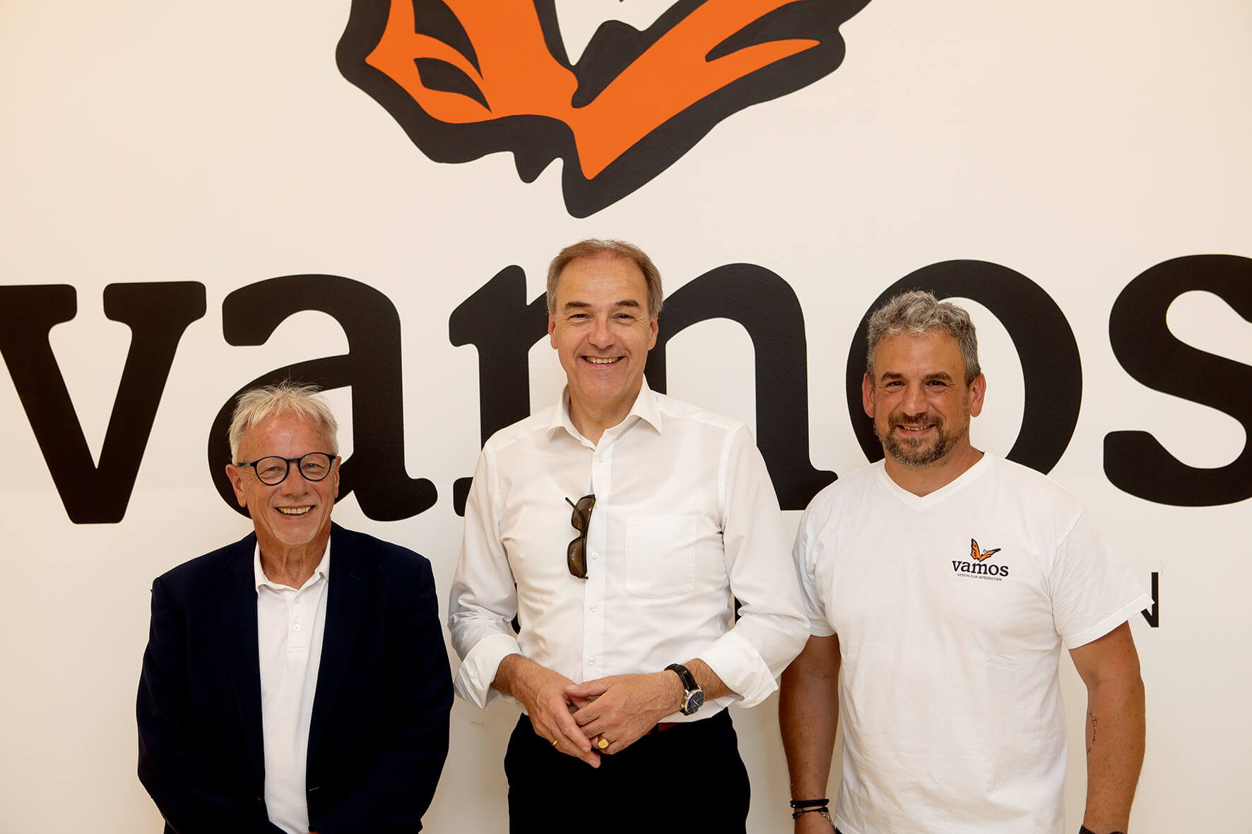 Drei Männer vor dem Vamos-Logo lächeln in die Kamera.