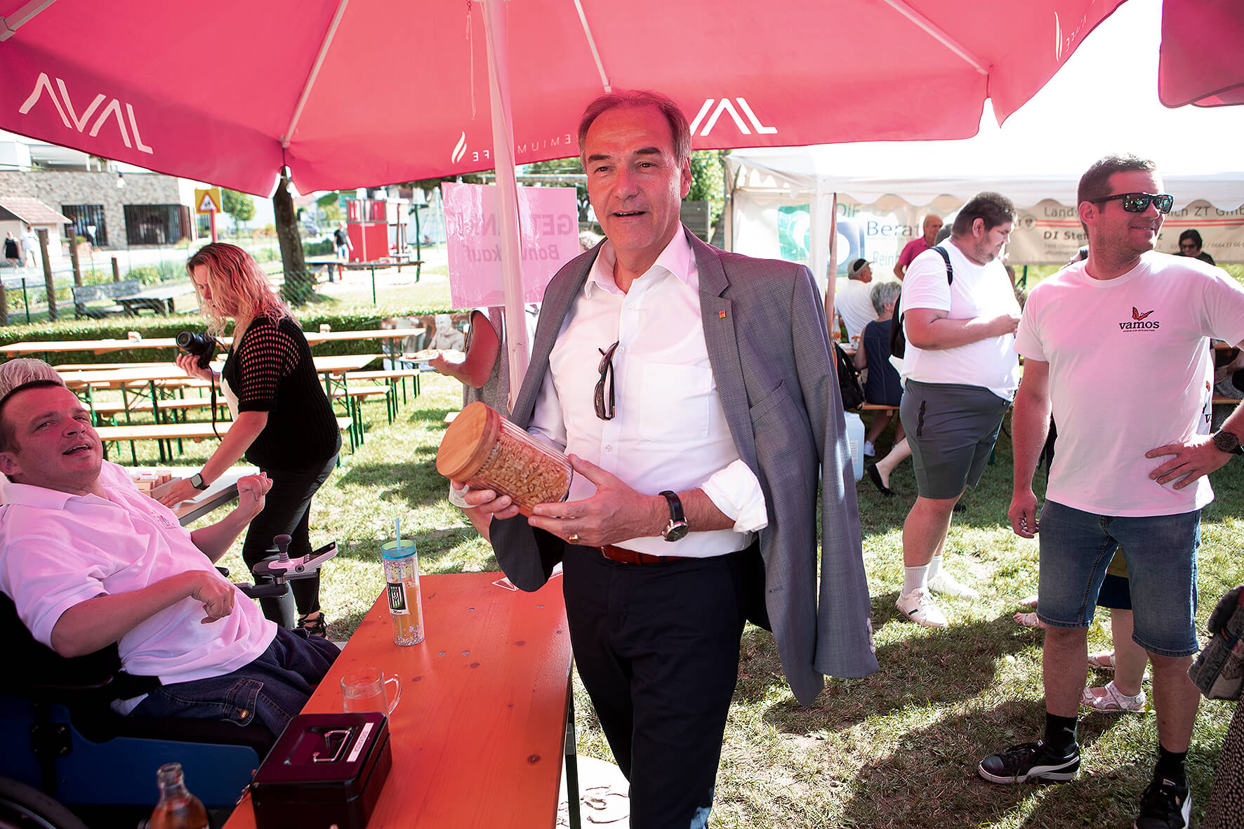 Mann mit Anzug lächelt unter pinkem Sonnenschirm bei Sommerfest im Freien und hält Glas mit Keksen in der Hand.