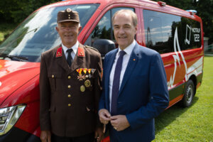 Zwei Männer vor einem Feuerwehrfahrzeug, einer in Uniform mit Medaillen, der andere im Anzug. Notruf 122 sichtbar.