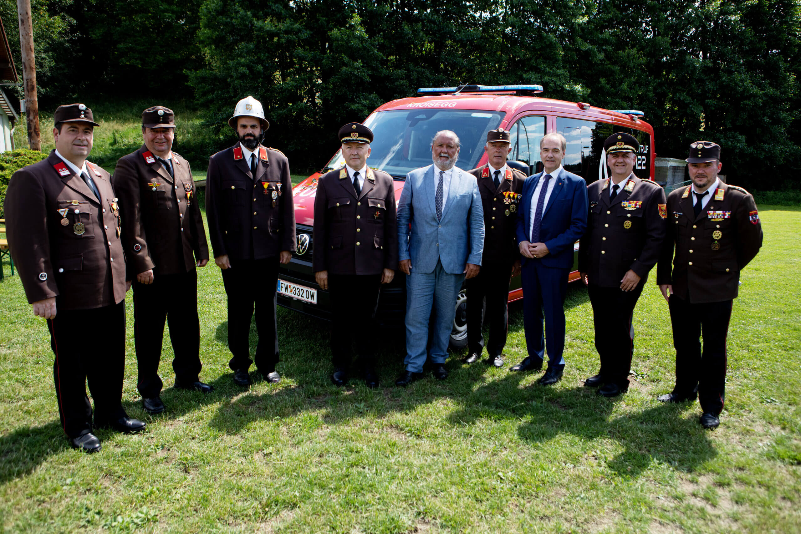Gruppe von Feuerwehrleuten in Uniform vor einem roten Einsatzfahrzeug bei sonnigem Wetter auf einer Wiese.