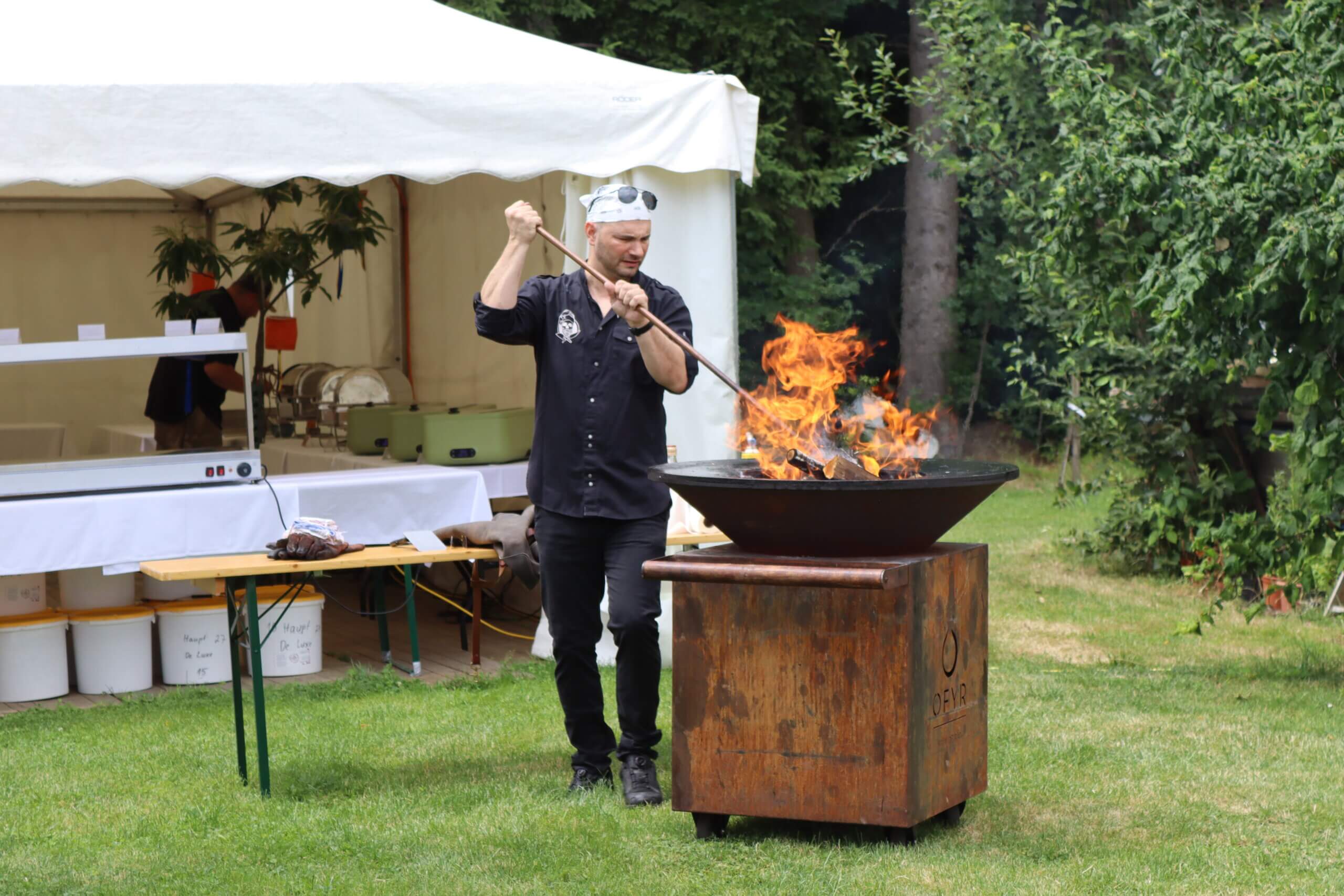 Outdoor-Koch bei Grillvorbereitung mit Feuerstelle, Catering-Zelt im Hintergrund, grüner Garten.