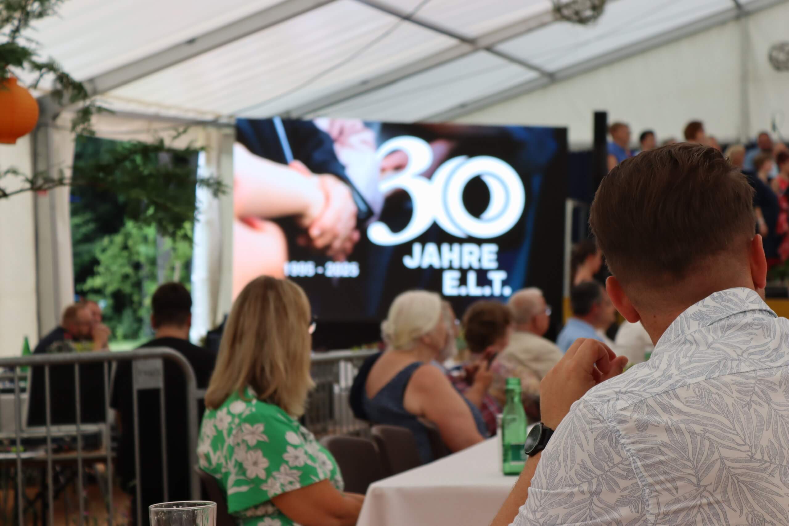 Feier zum 30-jährigen Jubiläum von E.L.T. mit Gästen in einem Festzelt, Leinwand im Hintergrund.