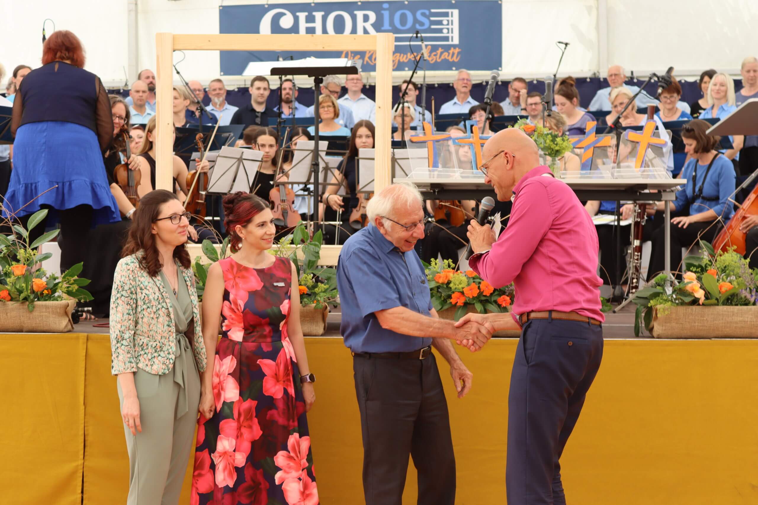 Menschen auf Bühne bei Konzert, Mann in pinkem Hemd schüttelt freundschaftlich einem älteren Herrn die Hand.
