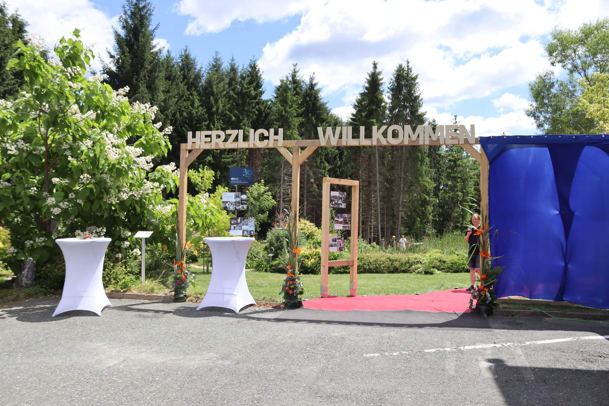 Herzlich Willkommen Schild bei Outdoor-Event mit dekorierten Stehtischen und Naturkulisse im Hintergrund.