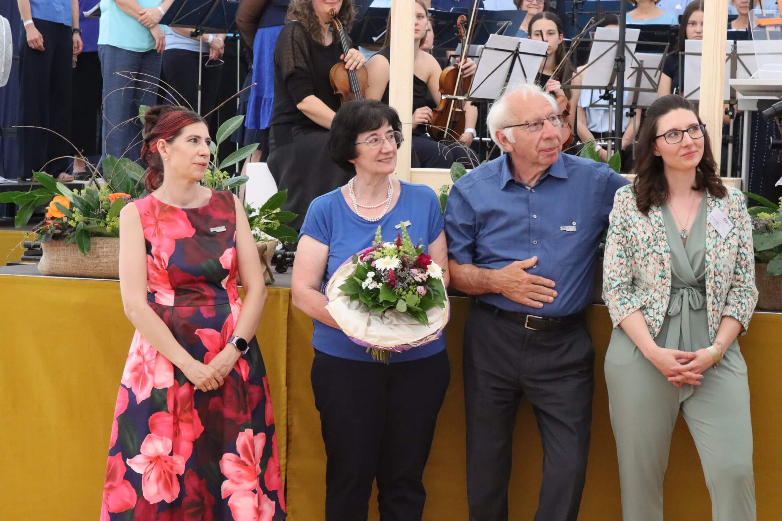 Personengruppe vor Orchester, Frau mit Blumenstrauß in der Mitte. Feierliche Veranstaltung mit festlicher Kleidung.