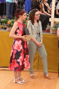 Zwei Frauen bei einem Preisverleih in eleganter Kleidung, eine hält eine Trophäe.
