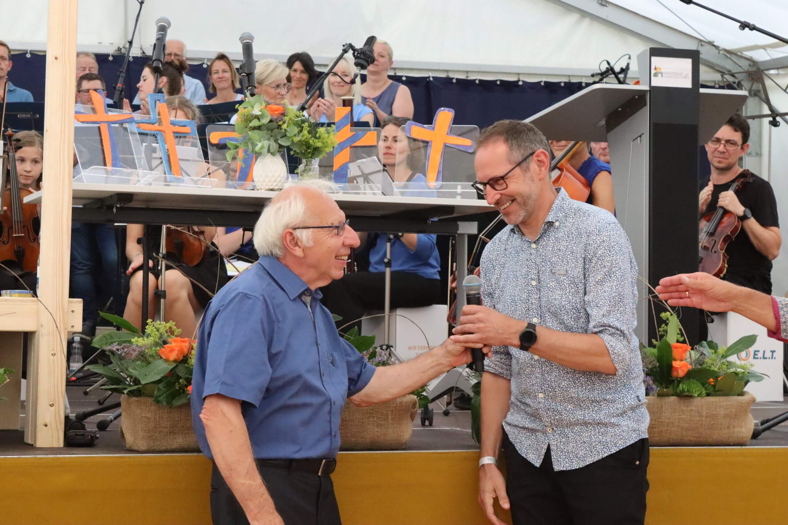 Älterer Mann und jüngerer Mann lächelnd bei einer Feier mit Musikern im Hintergrund auf einer Bühne.