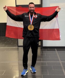 Person mit Medaille und Österreich-Flagge feiert Erfolg in Sporthalle.