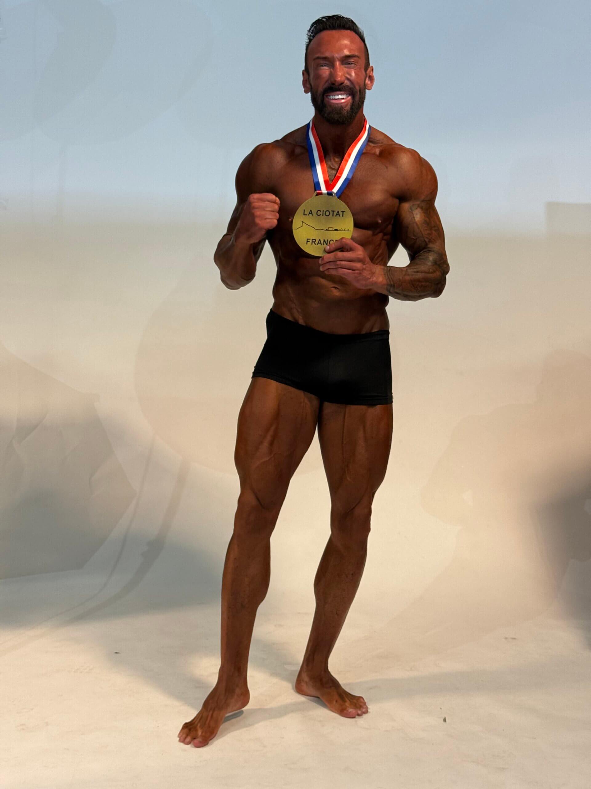 Bodybuilder mit Goldmedaille feiert Sieg in La Ciotat, Frankreich. Muskelkraft und Disziplin im Fokus.
