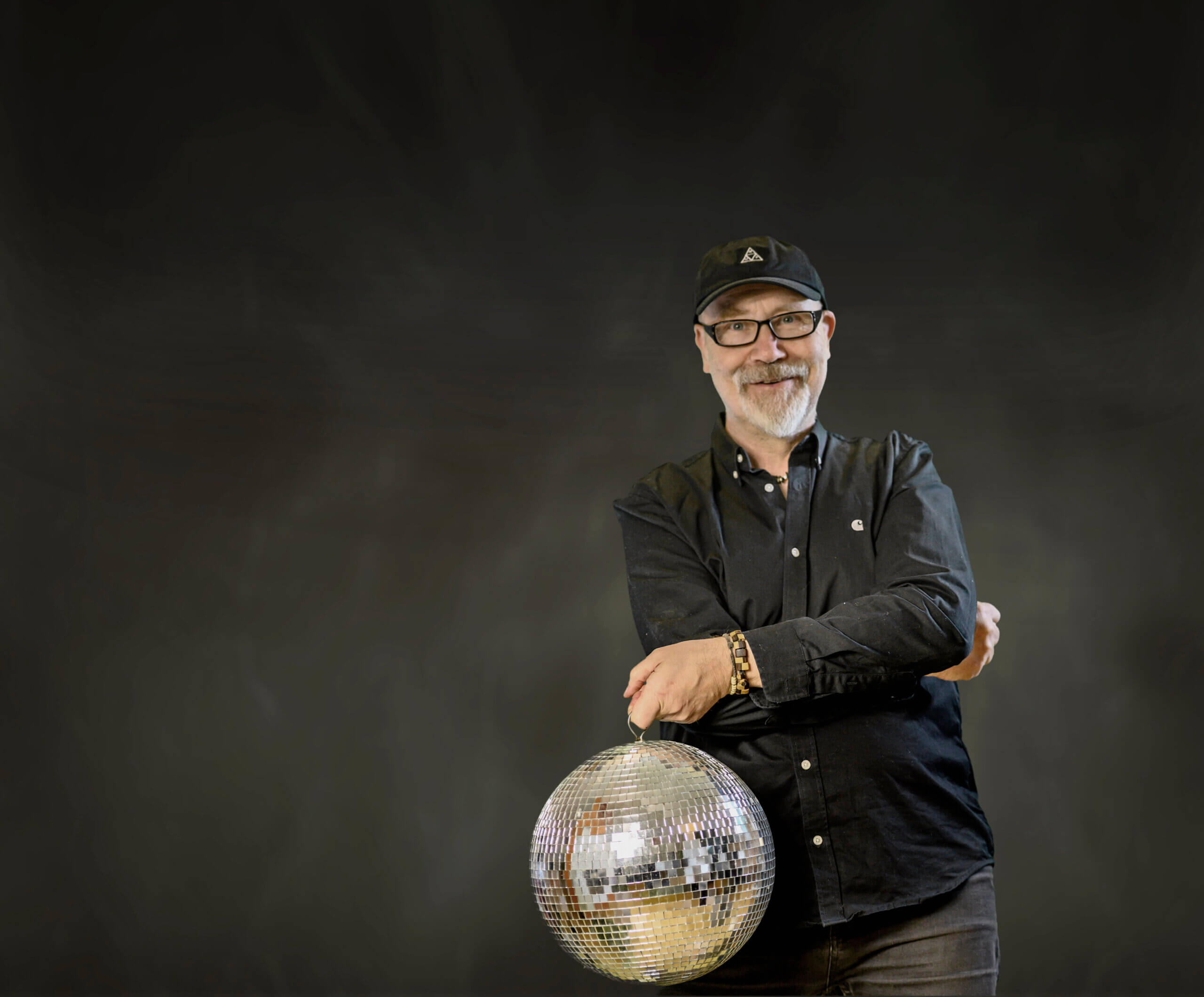 Mann in dunkler Kleidung hält eine silberne Diskokugel vor schwarzem Hintergrund.