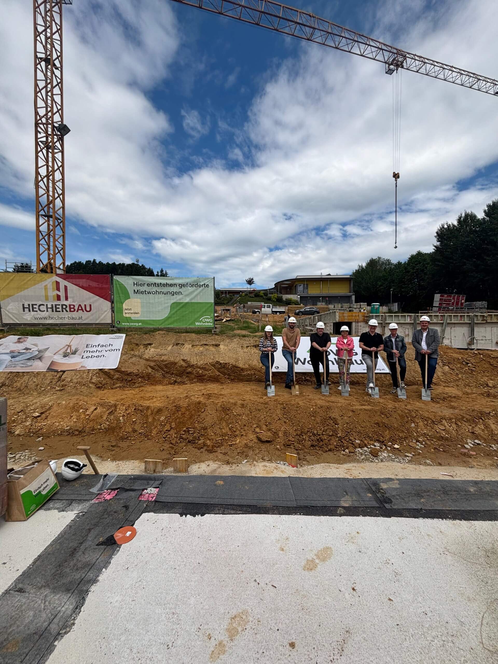 Personen beim Spatenstich auf einer Baustelle für geförderte Mietwohnungen, umgeben von Kran und Werbebanner.