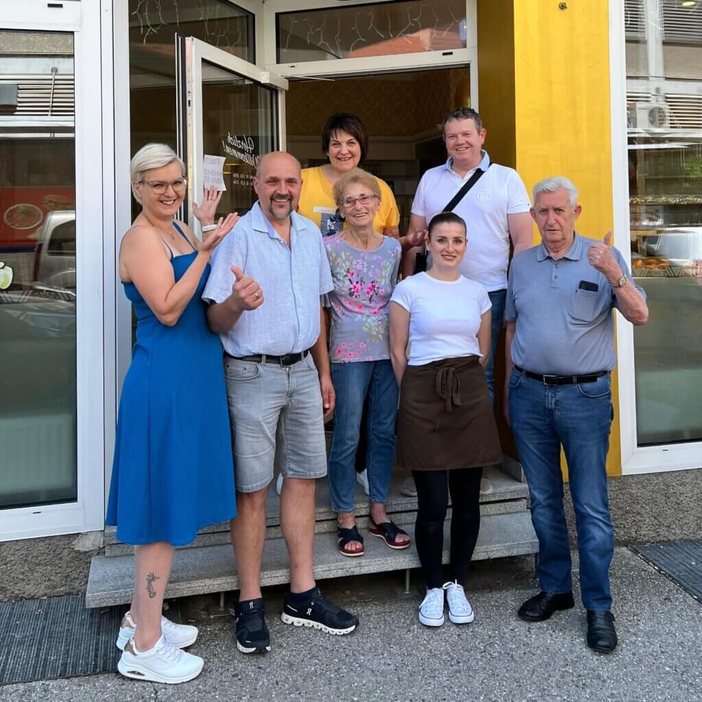 Gruppe fröhlicher Menschen vor einem Café-Eingang, Daumen hoch für einen gelungenen Tag.