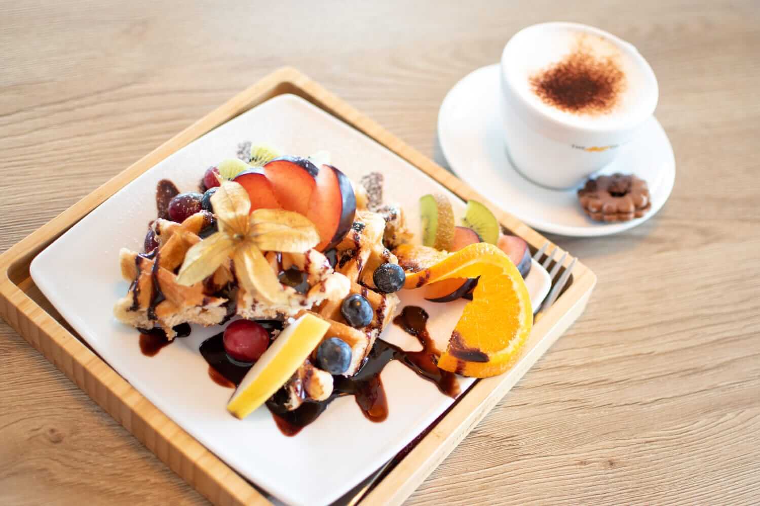 Waffeln mit frischen Früchten und Schokoladensoße auf einem Teller, dazu eine Tasse Cappuccino mit Keksen.
