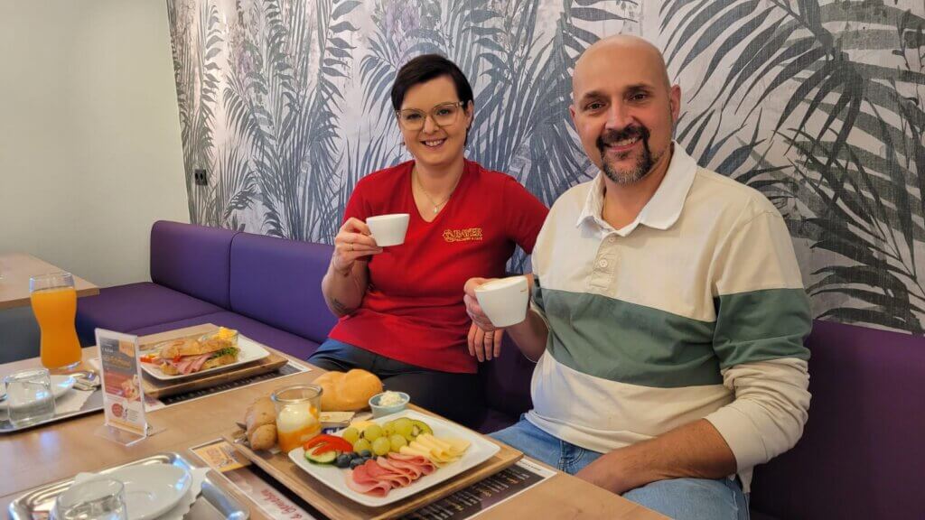 Paar genießt Frühstück mit Kaffee in einem Café, lächelnd vor einer tropischen Tapete.