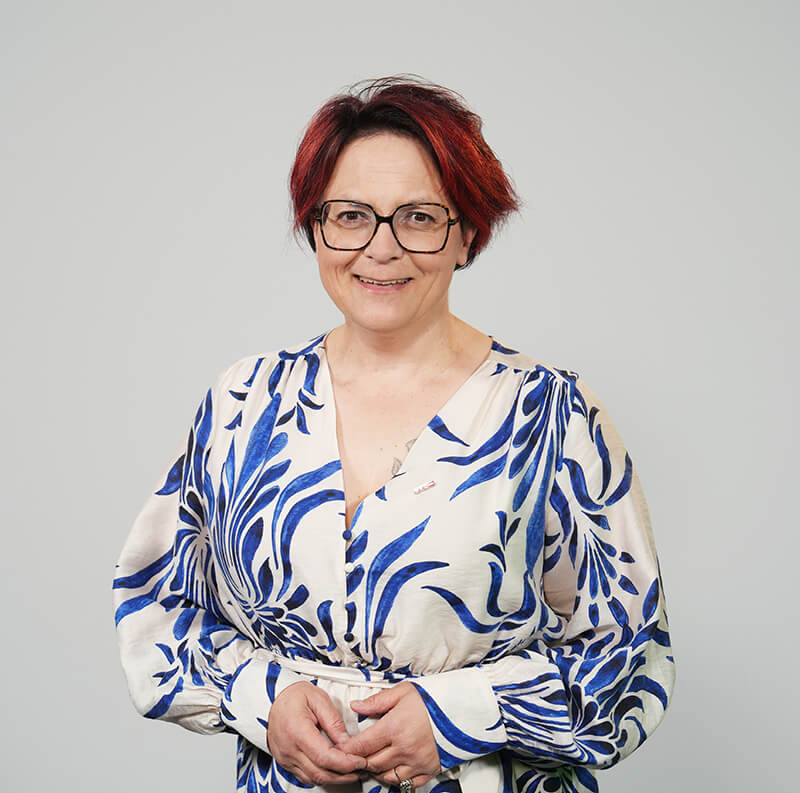 Tanja Stöckl©WK Oberwart Person mit Brille und rot schimmernden Haaren in einem weiß-blauen Kleid lächelt vor neutralem Hintergrund.