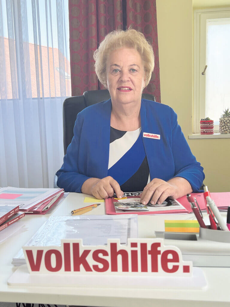 Verena Dunst©Volkshilfe Burgenland Eine ältere Frau sitzt an einem Schreibtisch mit Volkshilfe-Schild, umgeben von Büromaterialien und Unterlagen.