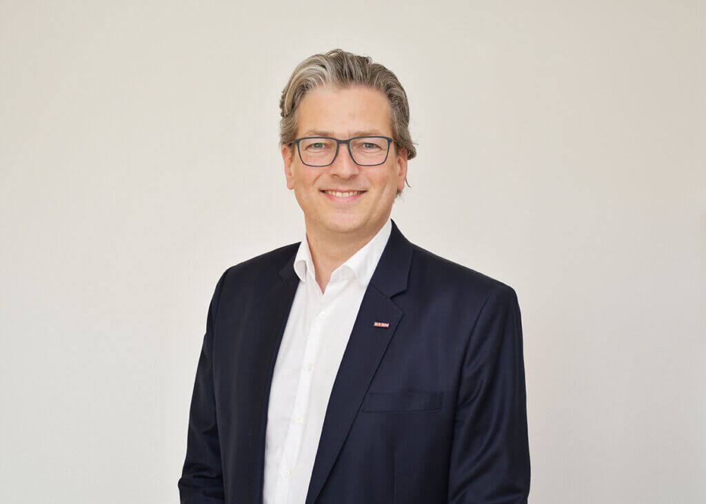 Winkler©WK Oberwart Eine lächelnde Person in Anzug und Brille vor neutralem Hintergrund.